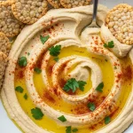 A creamy bowl of homemade gluten-free hummus drizzled with olive oil and sprinkled with paprika, surrounded by crunchy rice crackers partially dipped in the hummus on a rustic wooden board.